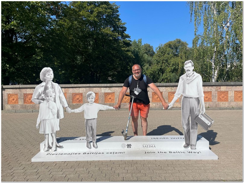 Eine Installation in Riga, die an den sog. Baltischen Weg erinnert. Auf ihr stehen vier fotorealistische Figuren in Schwarzweiß, die einander an den Händen halten. Links stehen eine Frau mit Kind, ein weiteres Kind, dann der Autor des Artikels, der wiederum Hand hält mit dem Mann auf der rechten Seite. So ist die Menschenkette interaktiv nachvollziehbar für alle.