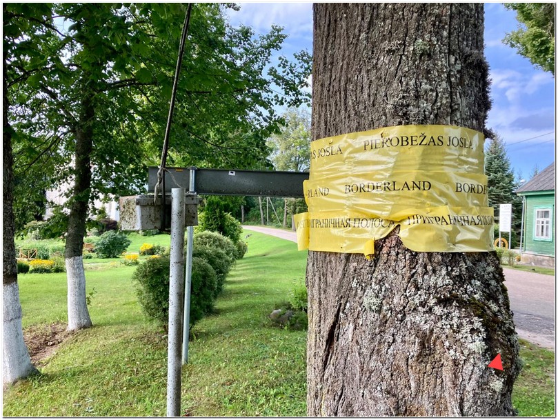 Ein Baum ist umwickelt mit einer gelben Markierungsfolie auf der Borderland (neben dem englischen Begriff stehen auch ein lettischer und ein russischer Text gleichen Inhalts auf dem Marker) steht. Das Bild ist aufgenommen in einem lettischen Grenzdorf an der Grenze von Lettland und Belarus/Weißrussland